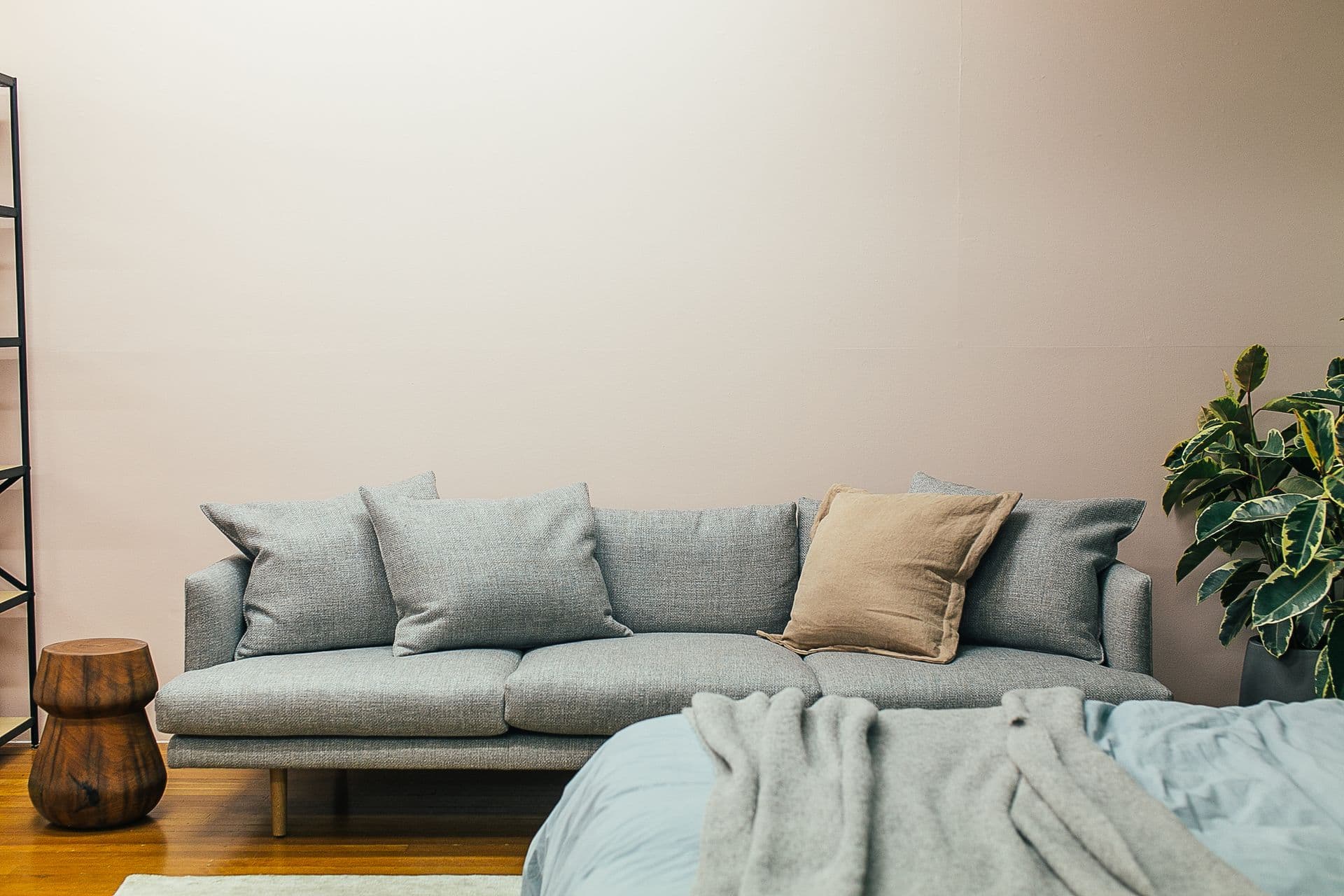 Un sofá gris claro con cojines contra una pared lisa beige, con un taburete de madera a un lado y una planta al otro.